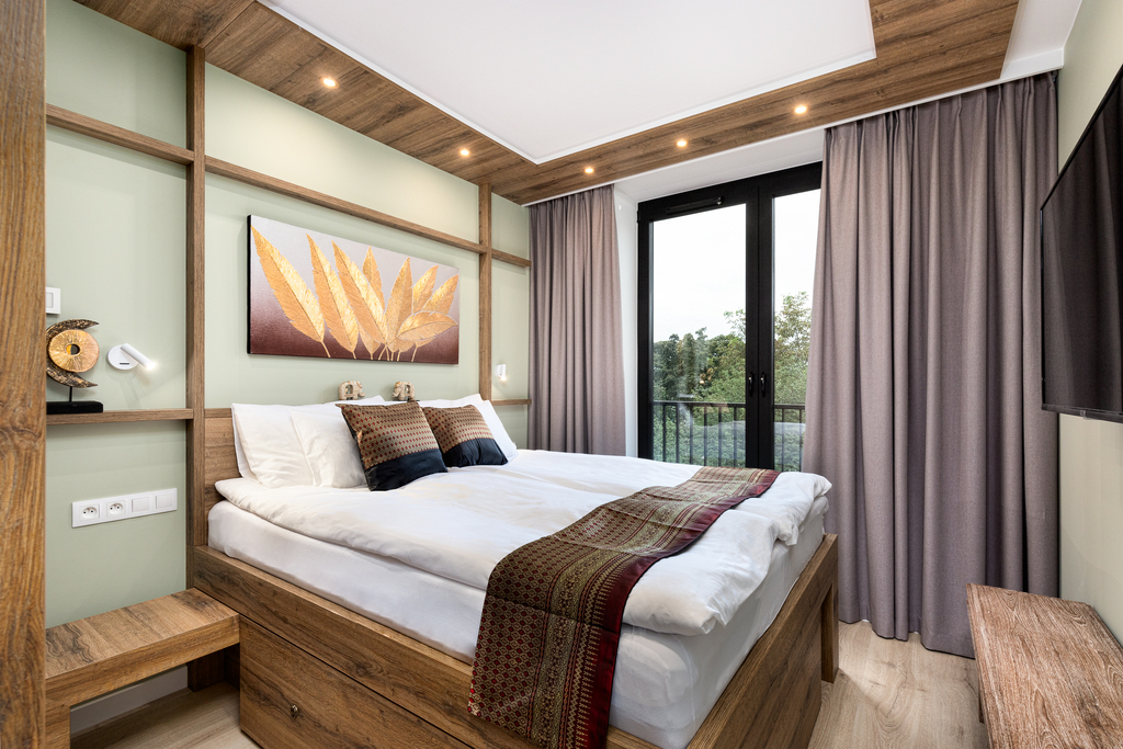 Schlafzimmer der Cosmea Suite mit Doppelbett, goldenem Blätterbild, Wand-TV und Balkontür ins Grüne